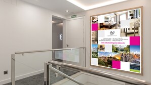 Pasillo de hotel con barandilla de vidrio, una puerta con la inscripción 'SALIDA' y un cartel publicitario del Grupo Woohoo con imágenes de atracciones de Madrid.