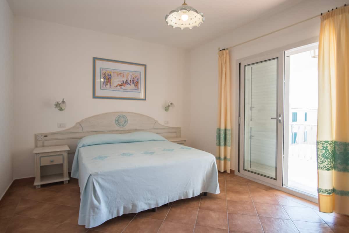 Un dormitorio con cama, mesita de noche, lámpara de pared, cuadro enmarcado y puerta corrediza de vidrio con cortinas.