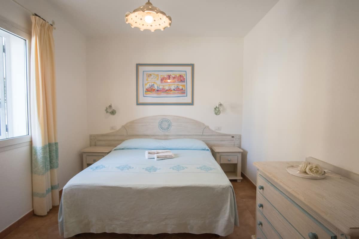 Un dormitorio con cama, mesita de noche, cómoda, ventana con cortinas e imagen colgada en la pared.