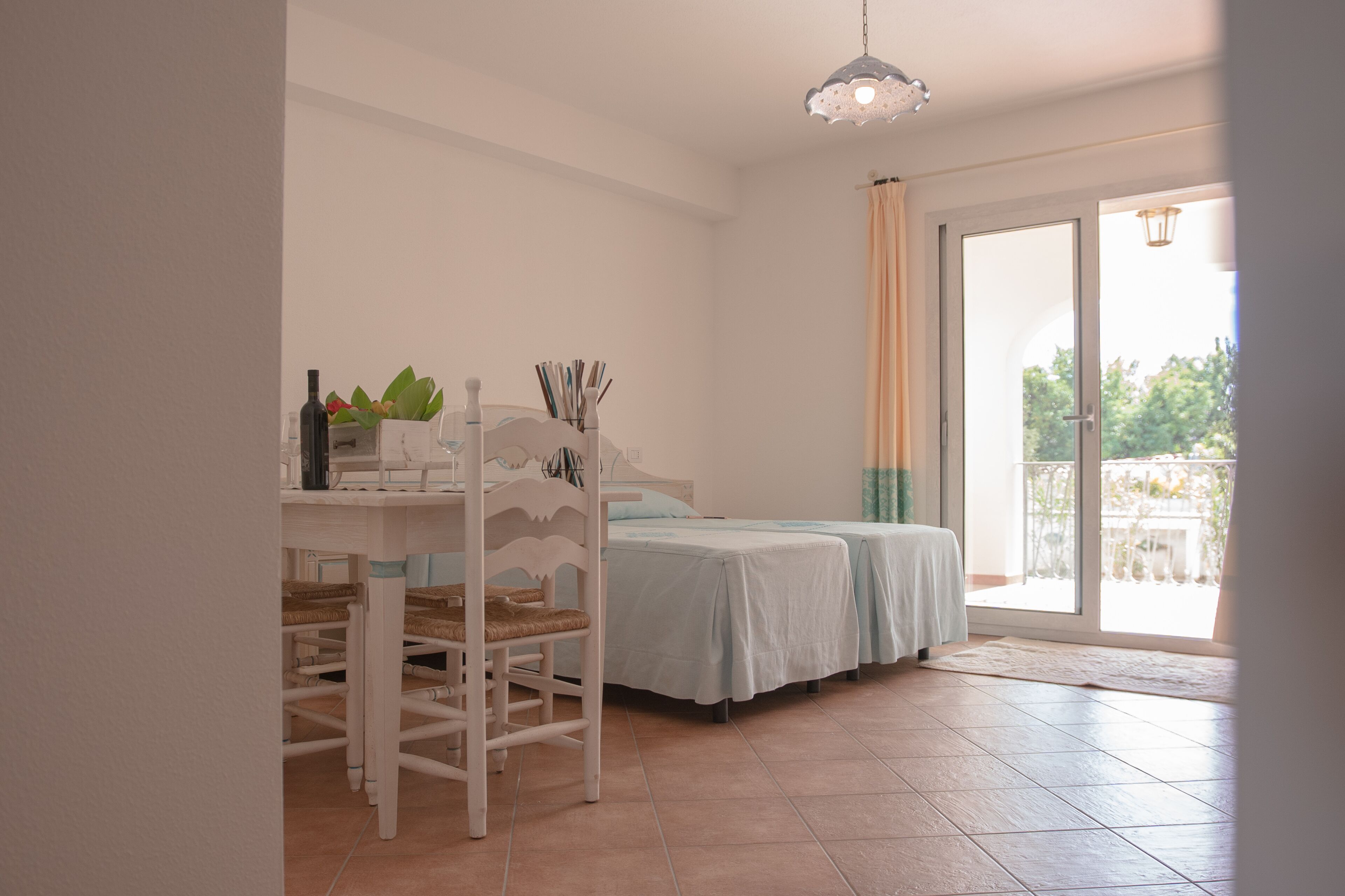A room with a bed, a table with chairs, a hanging light fixture, and a view through a glass door to an outdoor area.
