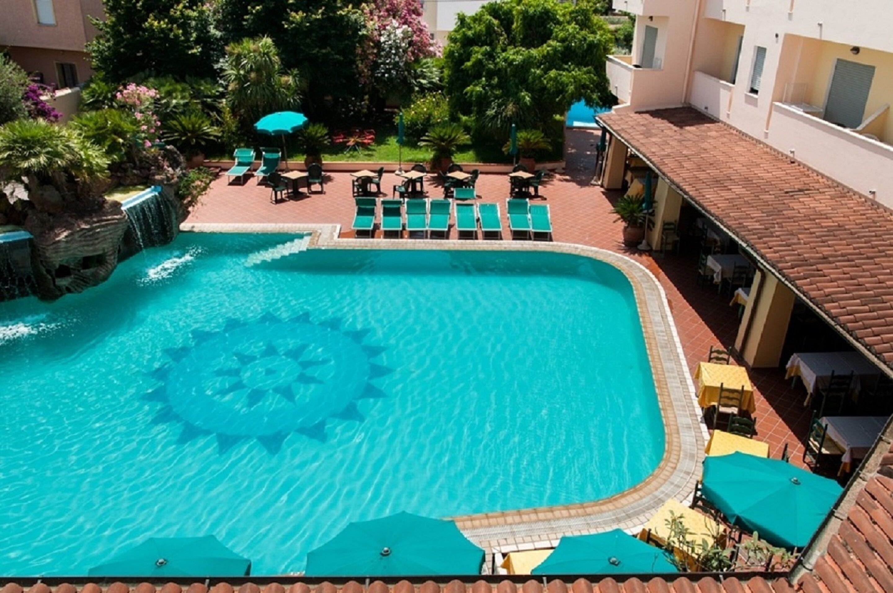 Una piscina de diseño circular, rodeada de sillones de playa y sombrillas, con una cascada y abundante vegetación de fondo.