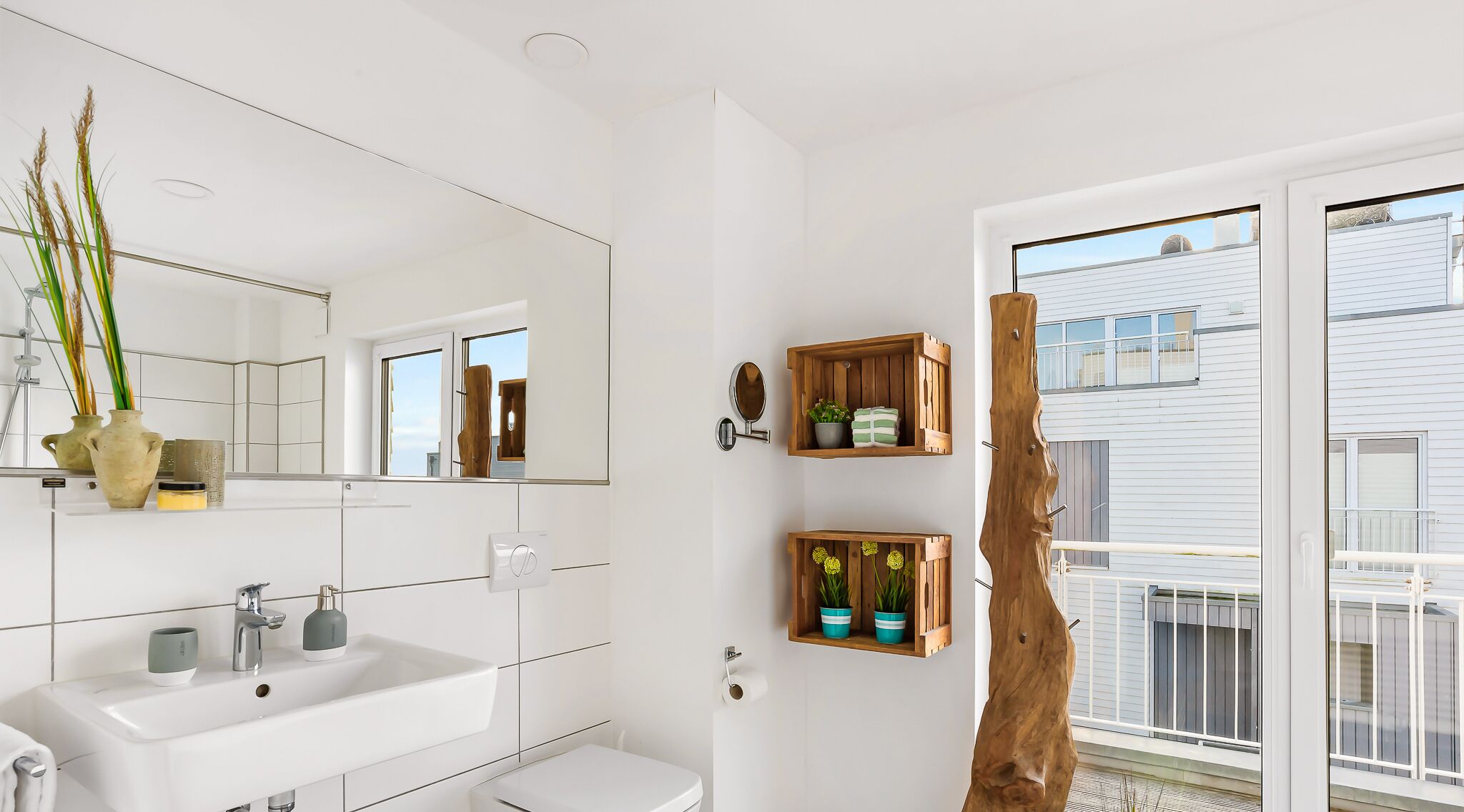 Une salle de bain moderne avec un grand miroir, des carreaux blancs, un lavabo et deux étagères en bois ornées de plantes.