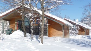 Una cabaña de madera con techo cubierto de nieve, rodeada de terreno y árboles nevados.