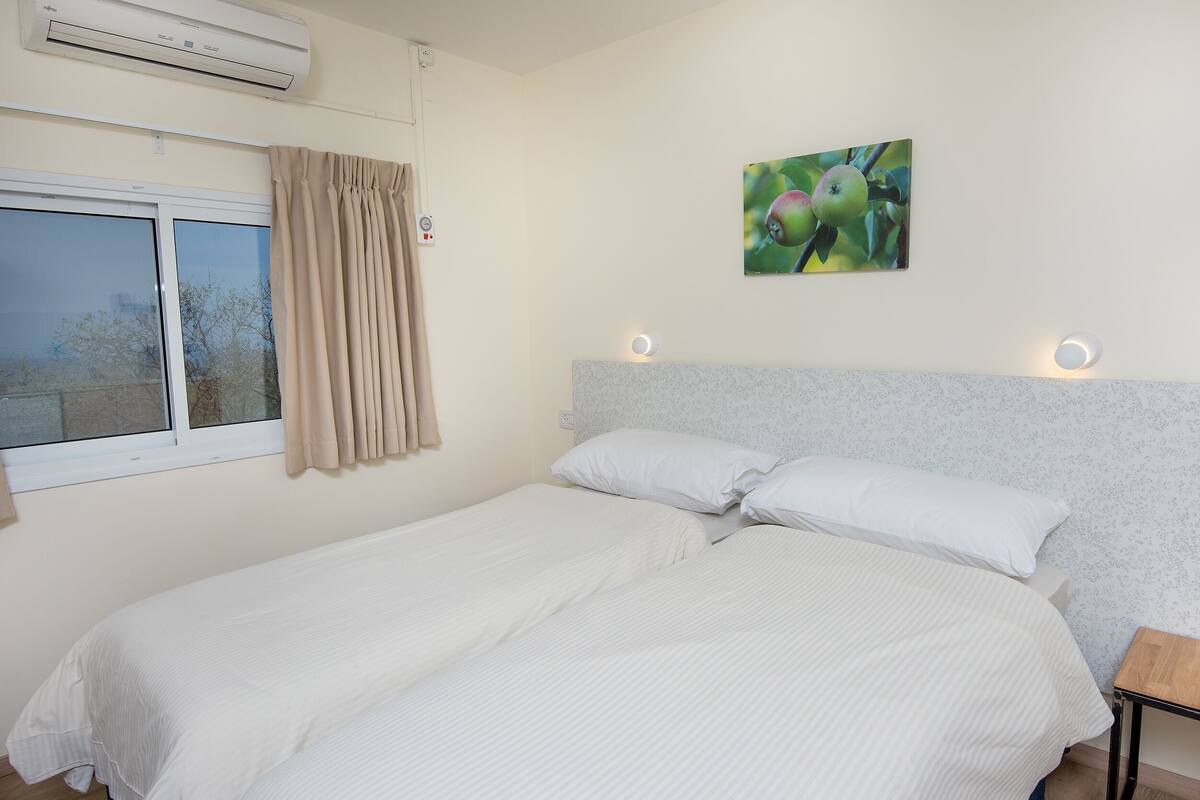 Un letto matrimoniale con biancheria da letto bianca, una testiera con illuminazione integrata, una finestra con tende e un quadro appeso al muro raffigurante della frutta.