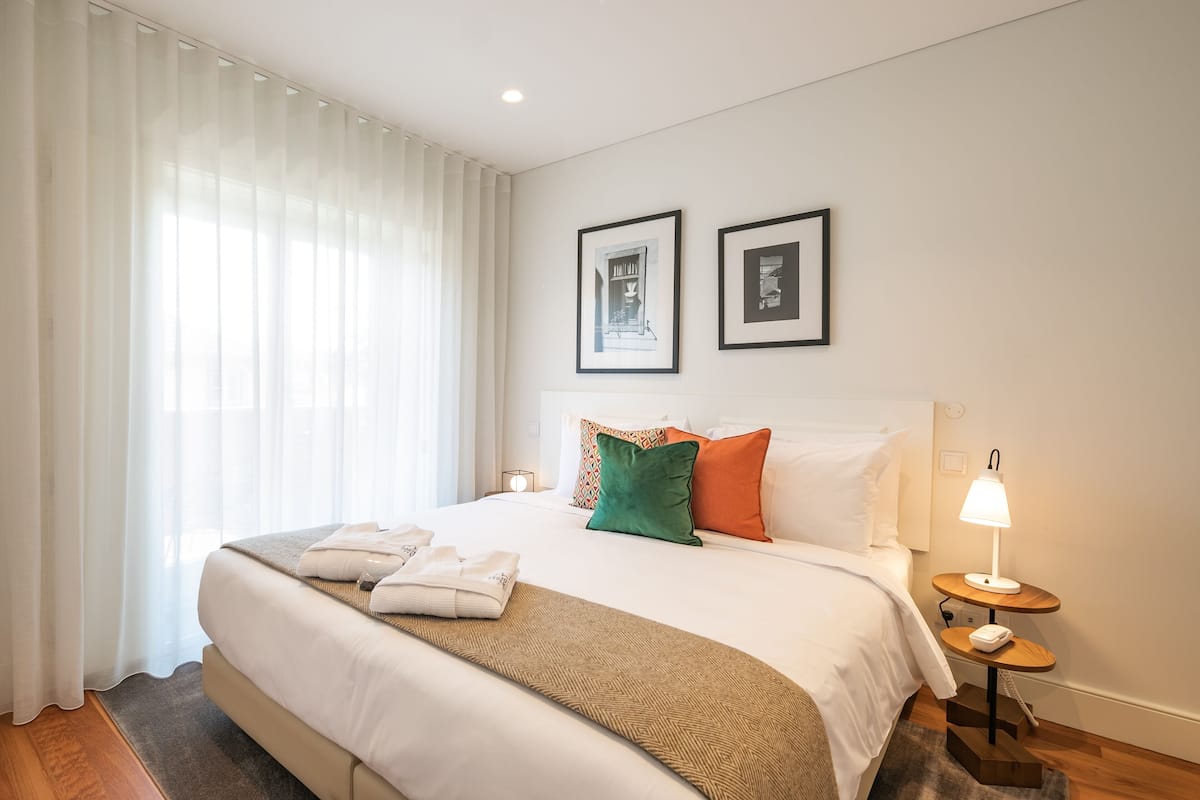 Una cama bien hecha con edredón blanco, una manta marrón y dos almohadas en verde y naranja. Hay una mesita de noche con una lámpara y una toalla doblada. Dos cuadros enmarcados están colgados en la pared sobre la cama.