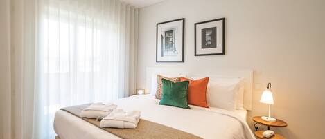 A neatly made bed with a white duvet, a brown throw blanket, and two pillows in green and orange. There is a small bedside table with a lamp and a folded towel. Two framed pictures are hung on the wall above the bed.