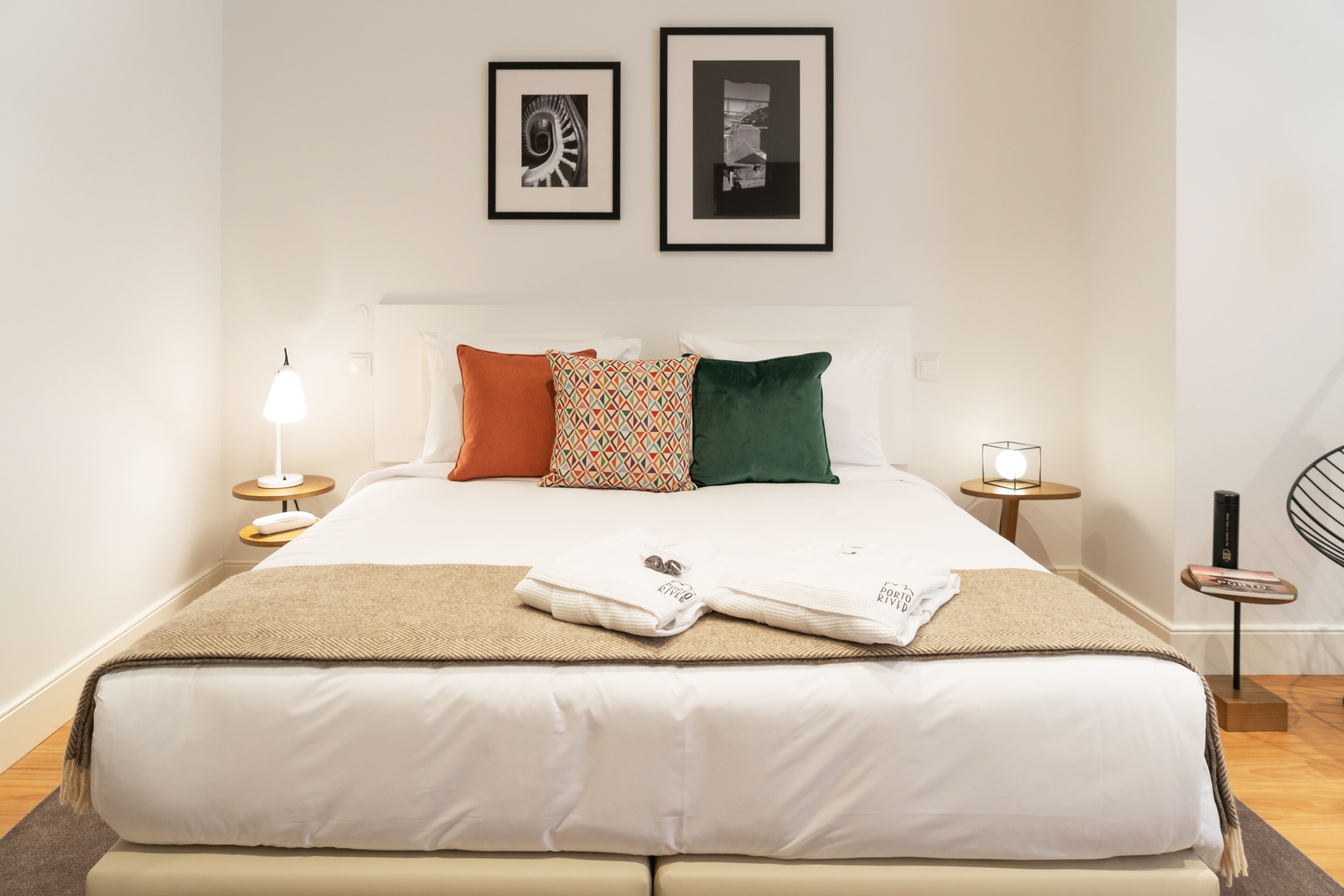 Un lit bien fait, recouvert d’une couette blanche, agrémenté d’un coussin à motifs et d’un coussin vert. Deux tables de chevet avec des lampes se trouvent de chaque côté du lit, et un tableau encadré est accroché au-dessus.
