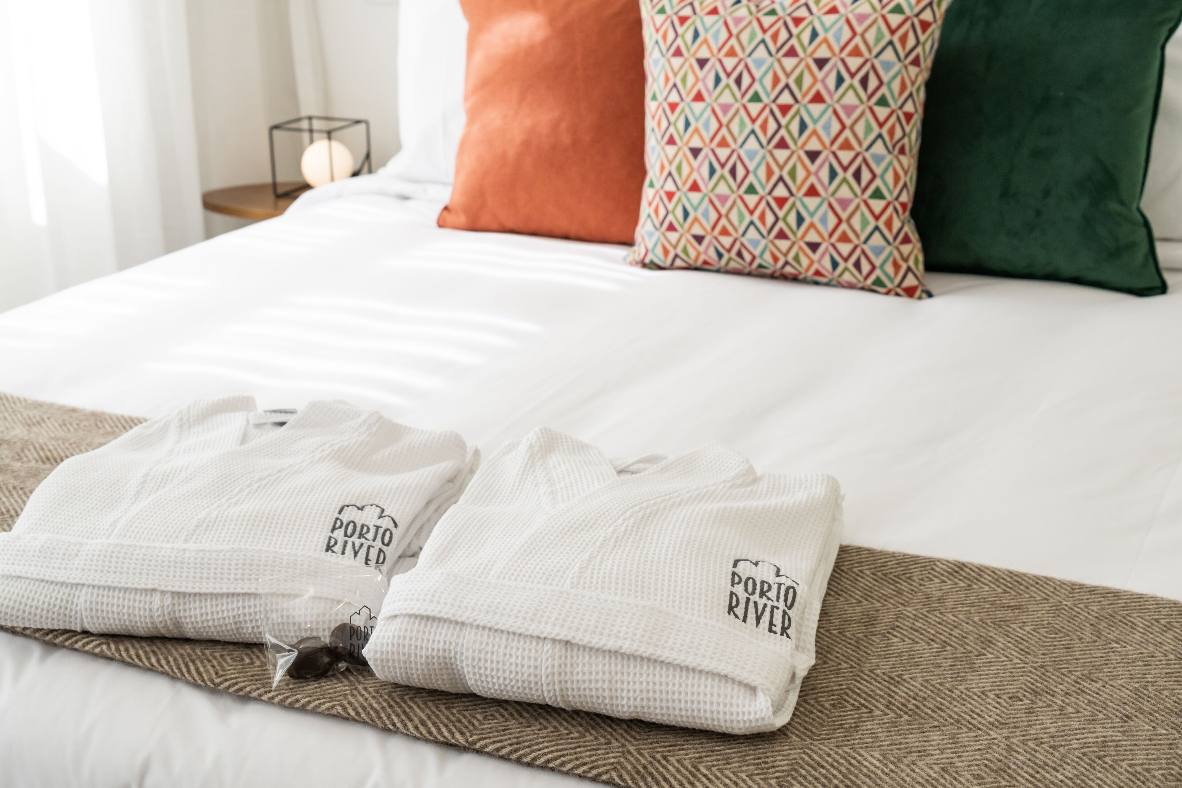 A neatly made bed with white linens, two folded towels with 'Porto RIVIER' branding, and a patterned pillow.