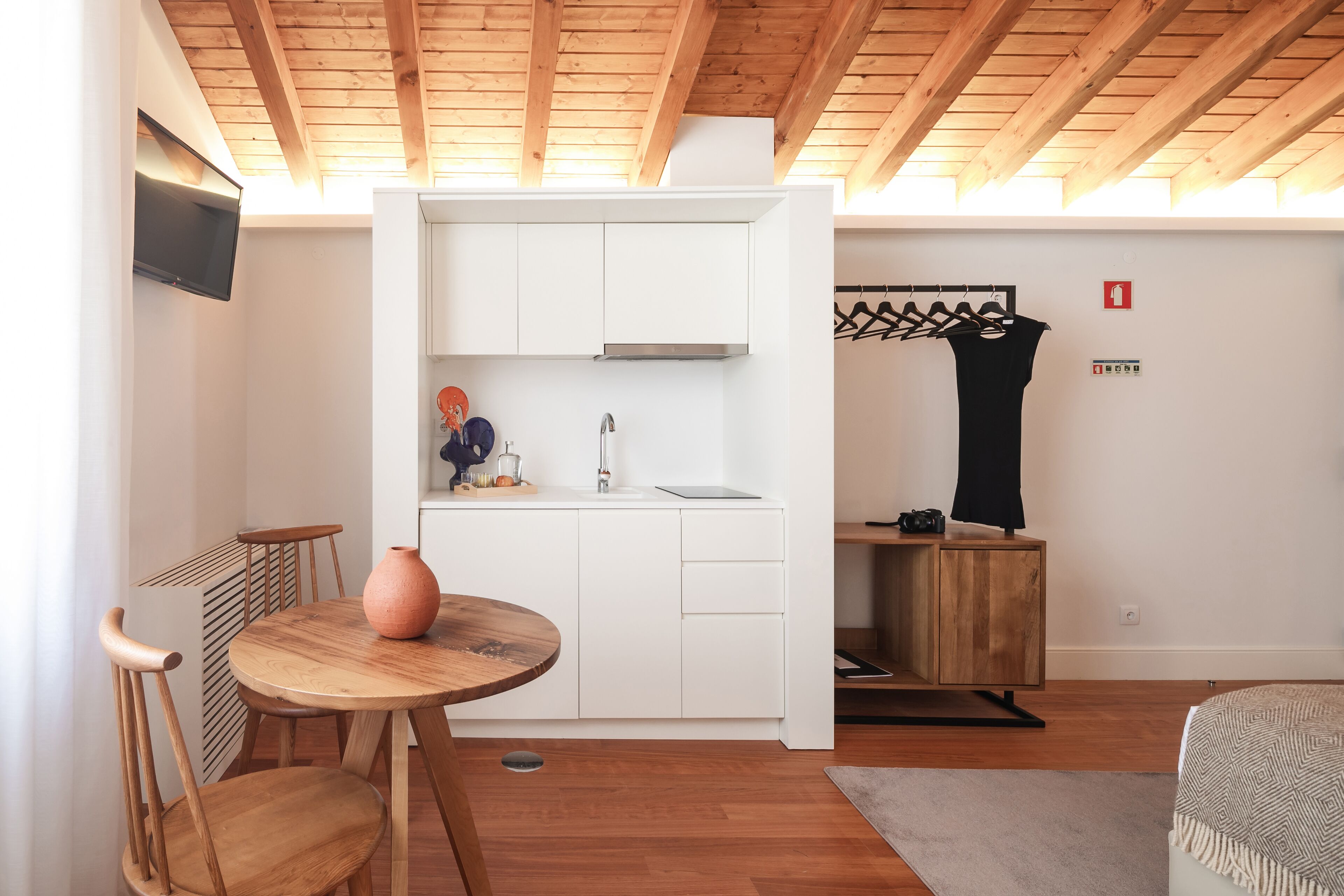 A compact kitchen with white cabinets, a wooden ceiling, a small dining table with chairs, and a hanging rack.