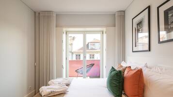 Una habitación de hotel con cama, almohadas, una silla y vistas a edificios a través de una puerta.
