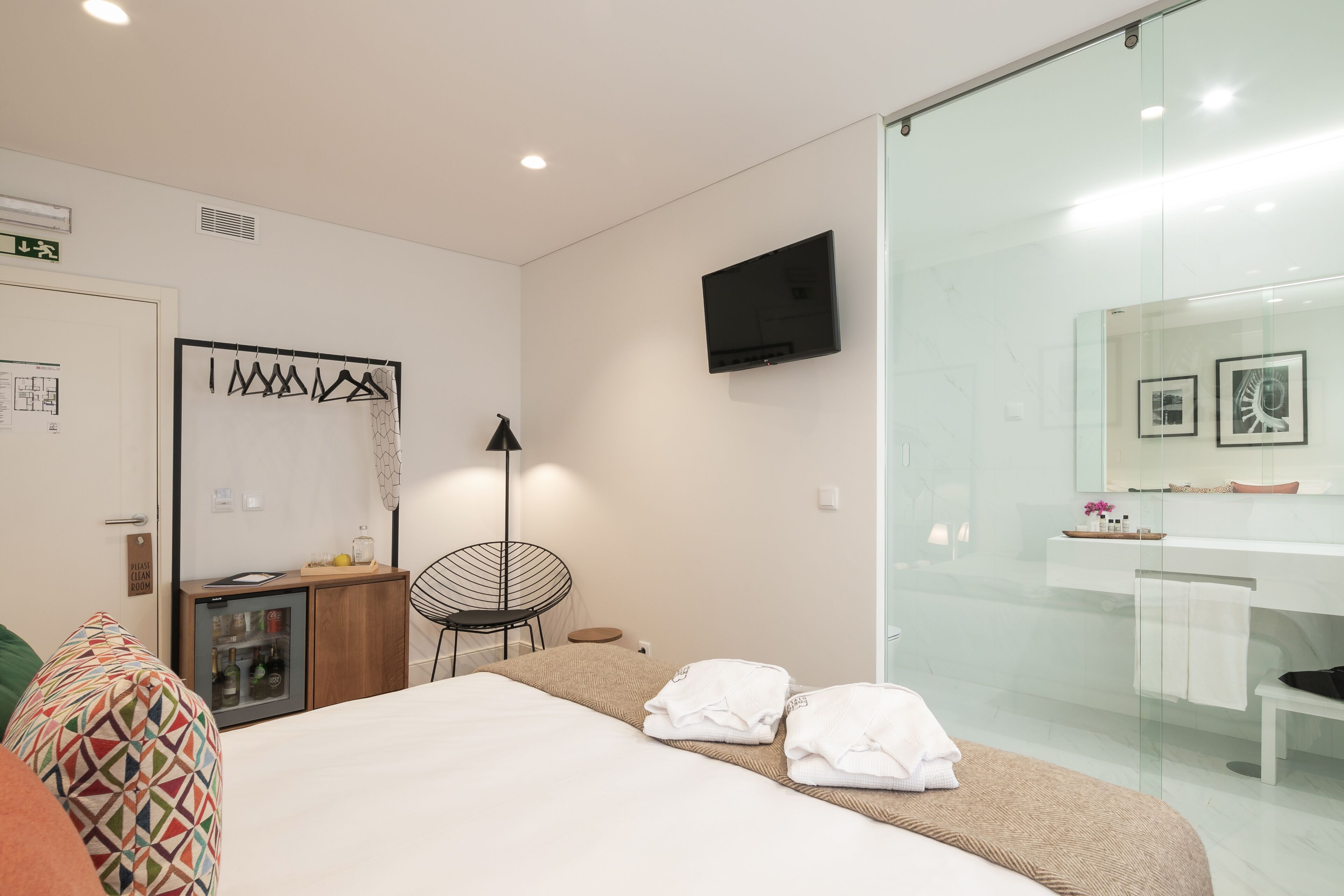 A modern hotel room with a bed, a small desk with a refrigerator, a chair, a TV mounted on the wall, and a bathroom with a glass shower.