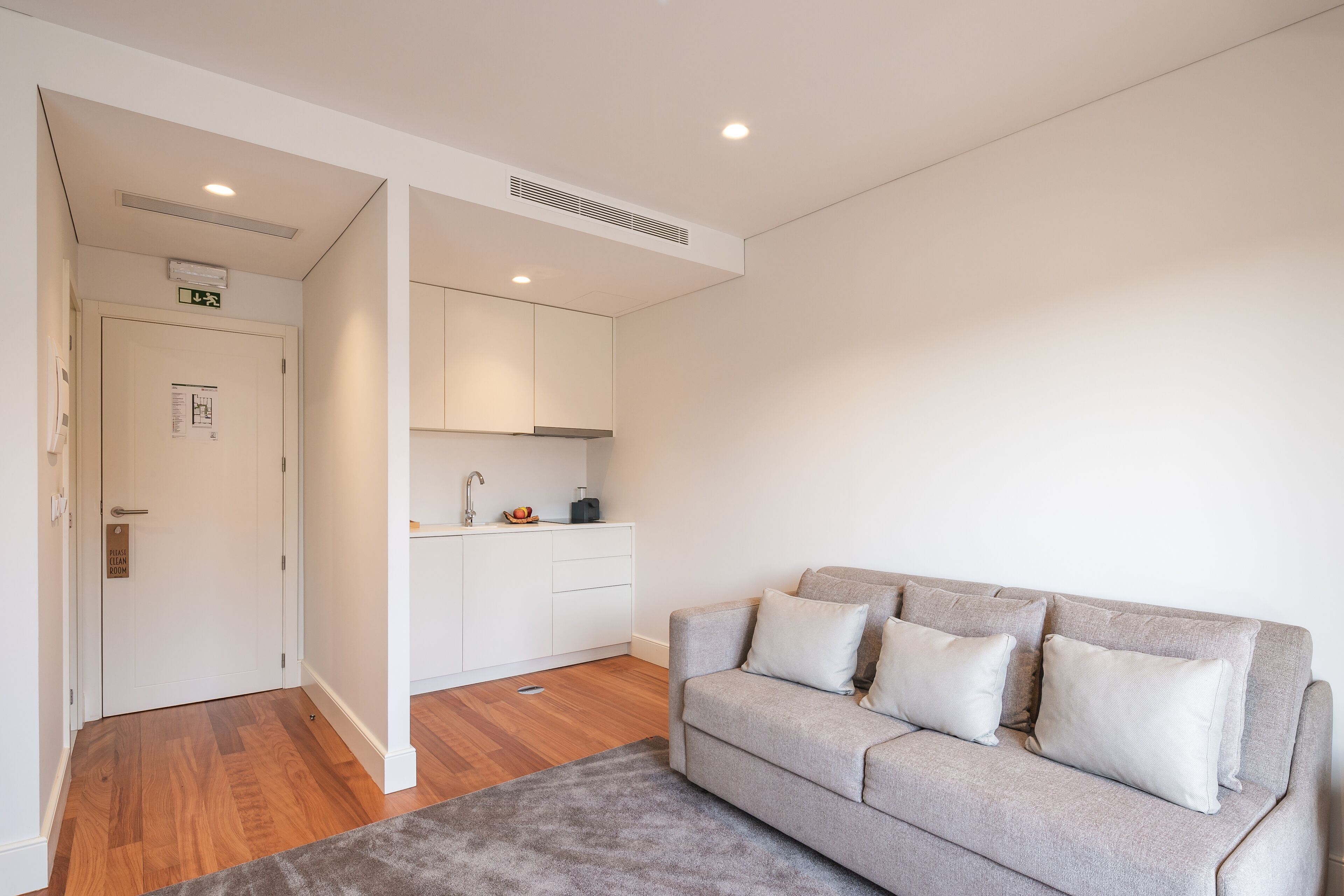A modern living room with a grey sofa, a small kitchen area, and a wooden floor.