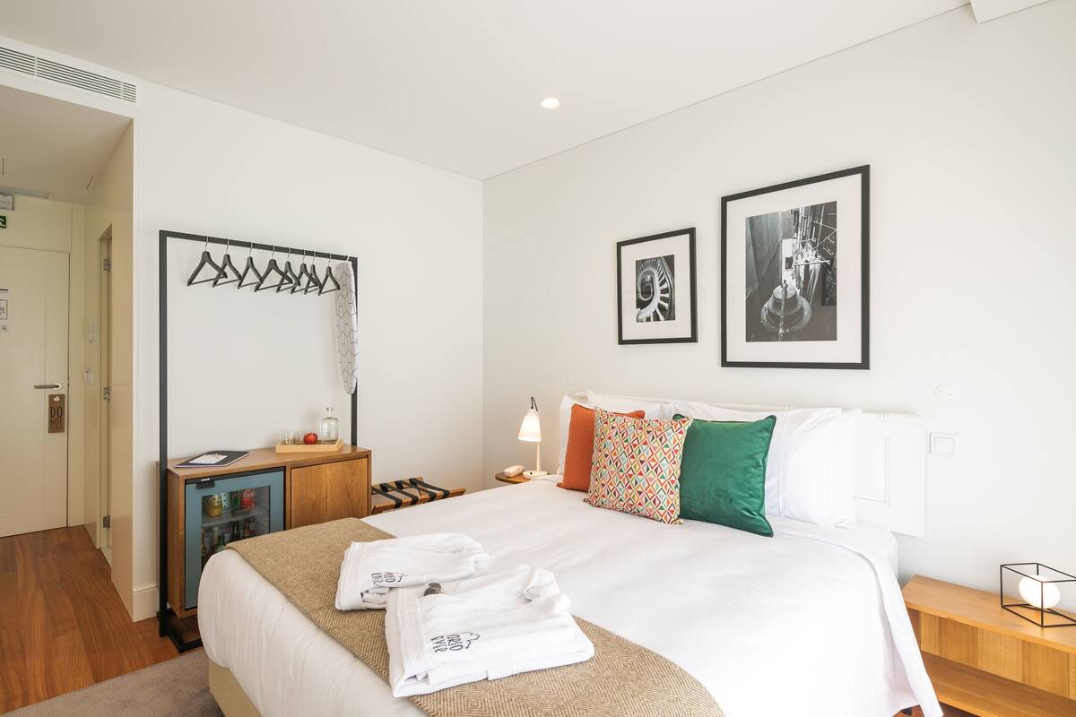 Habitación de hotel con una cama grande, una mesita de noche de madera, un escritorio con refrigerador y dos cuadros en la pared.