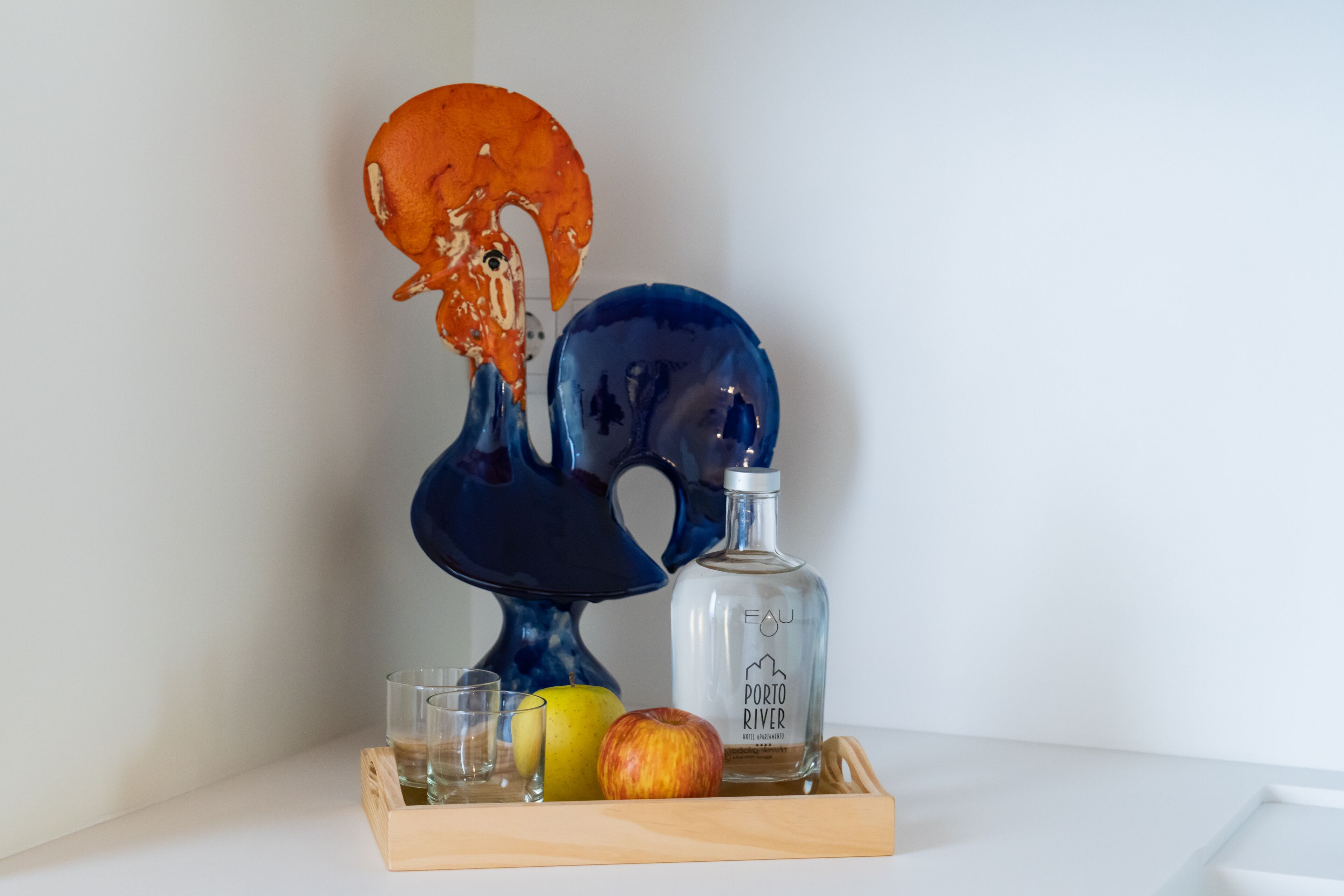 A decorative ceramic piece, a bottle labeled "Eau Porto River," and two glasses on a wooden tray with an apple.
