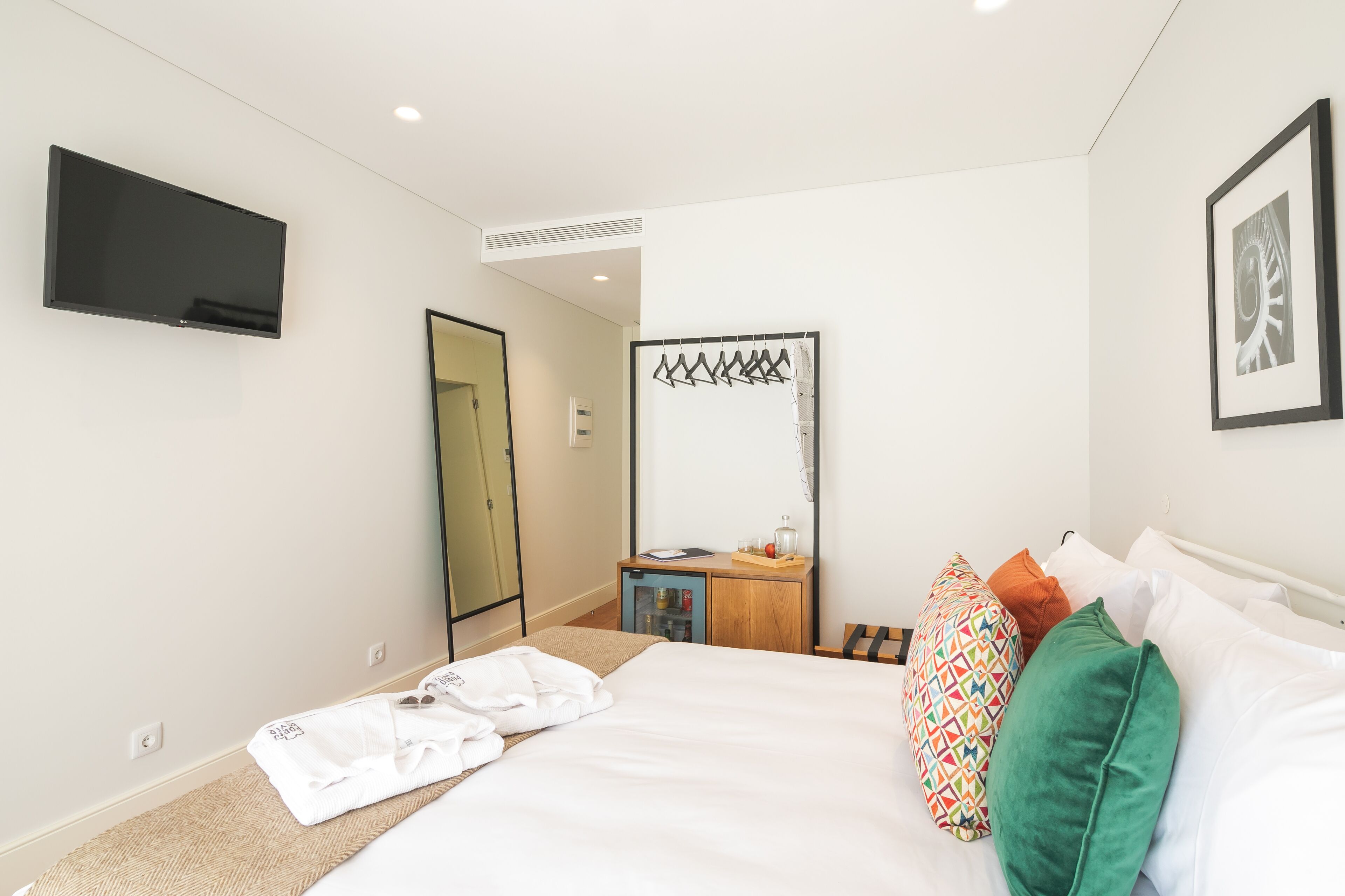 Una habitación de hotel con una cama, un televisor fijado a la pared, una mesita de noche de madera y un espejo.
