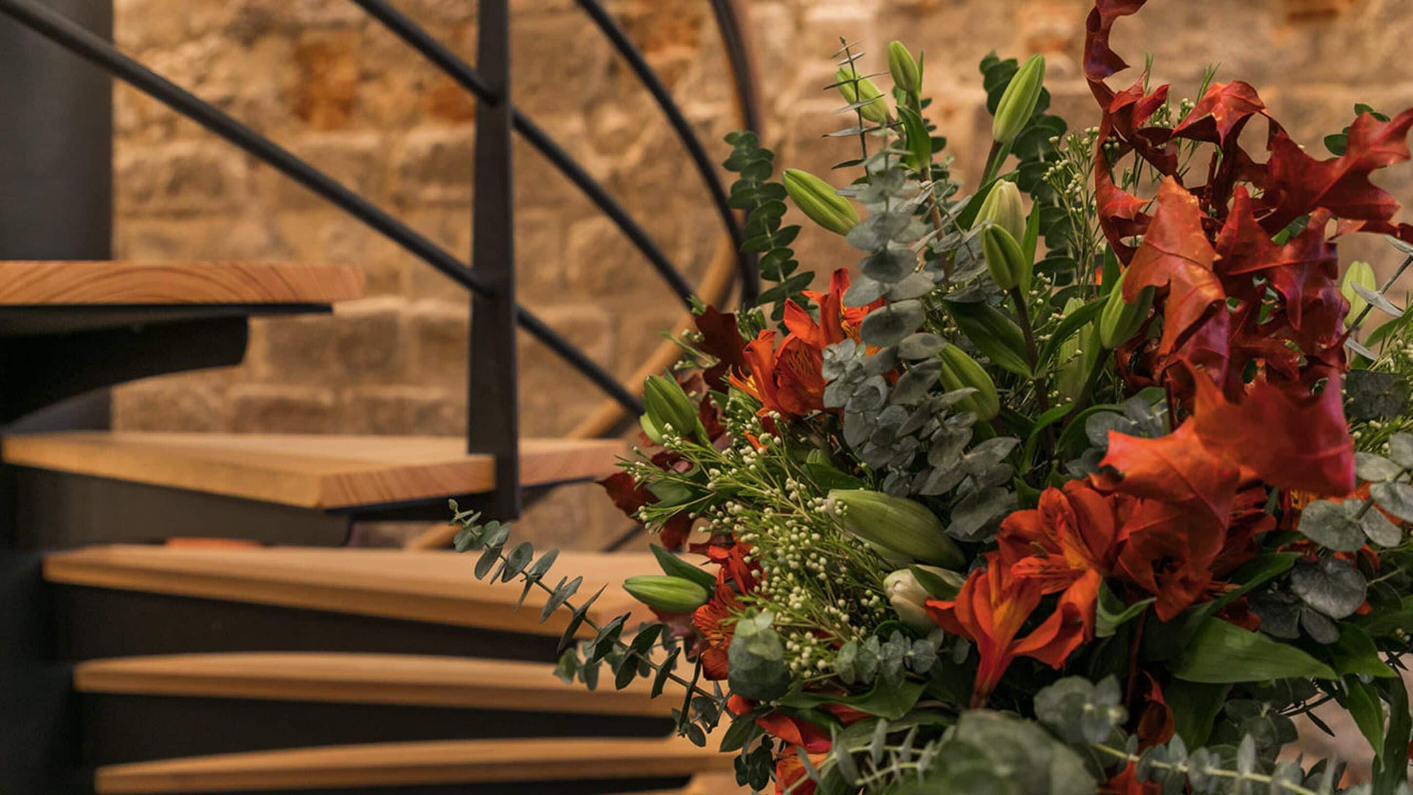 A floral arrangement with red and green foliage, placed on a wooden table against a stone wall background.