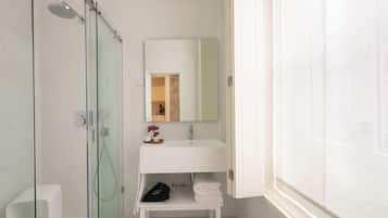 Un baño moderno con cabina de ducha de vidrio, un lavabo blanco con espejo y un estante pequeño con toallas y artículos de aseo.