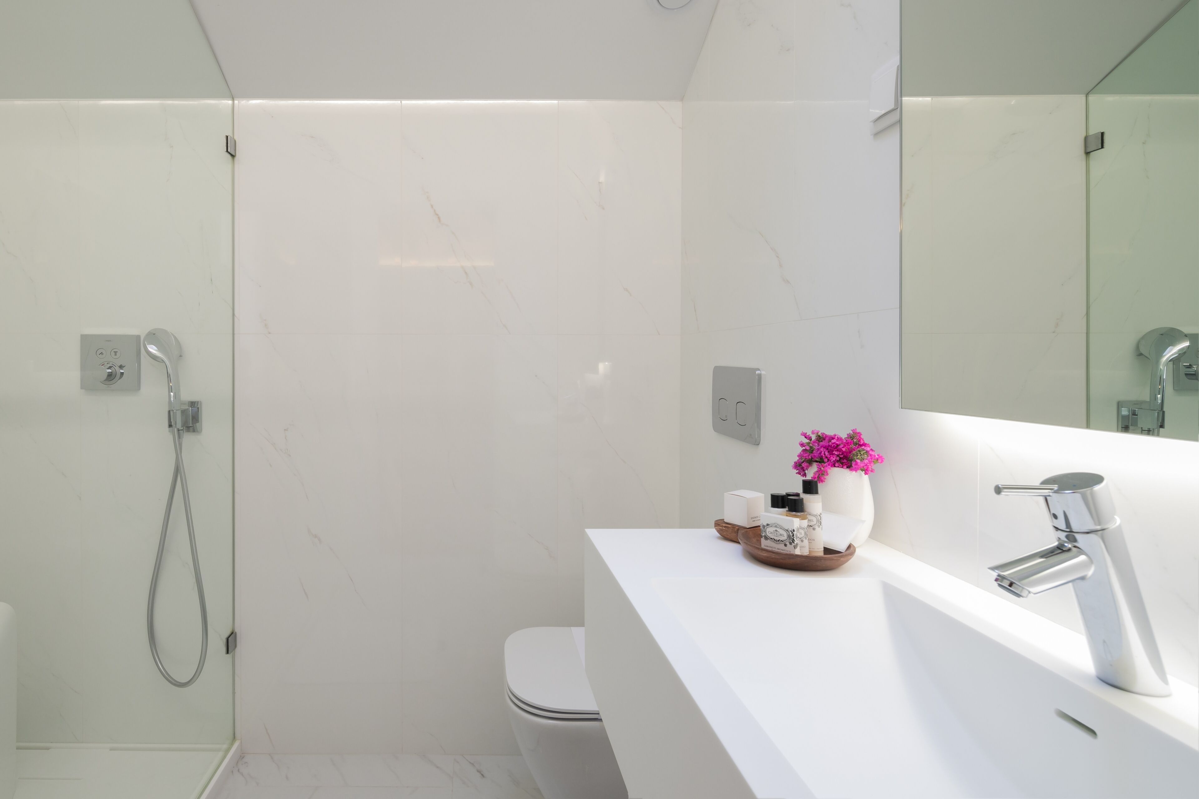 Une salle de bain moderne avec un mur en marbre blanc, une douche en verre, des toilettes blanches et un meuble-lavabo avec un miroir et un évier.