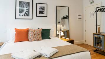 Una cama bien hecha con sábanas blancas, una almohada estampada y otra almohada verde. En la pared hay dos cuadros, un espejo y una mesita de noche con una lámpara y un teléfono.