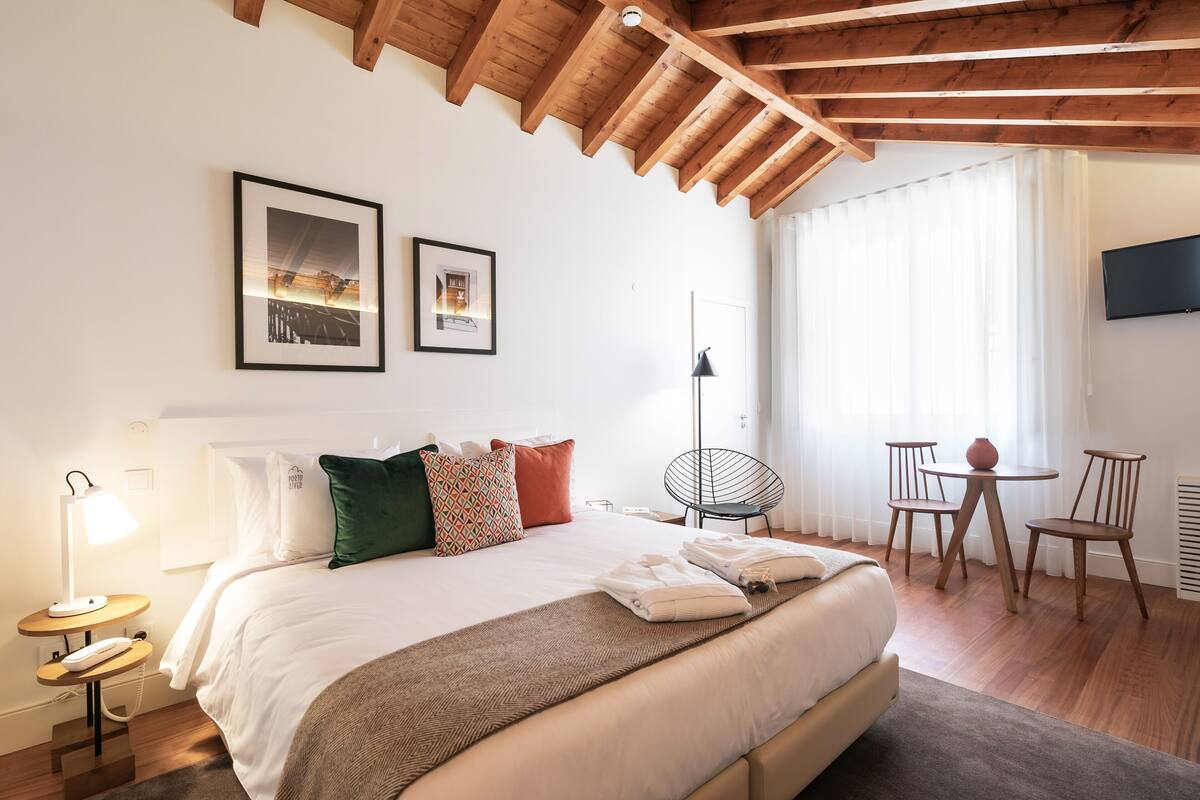 Un dormitorio con una cama grande, una mesita de noche con una lámpara, una silla, una pequeña mesa con un florero, y un televisor en la pared.