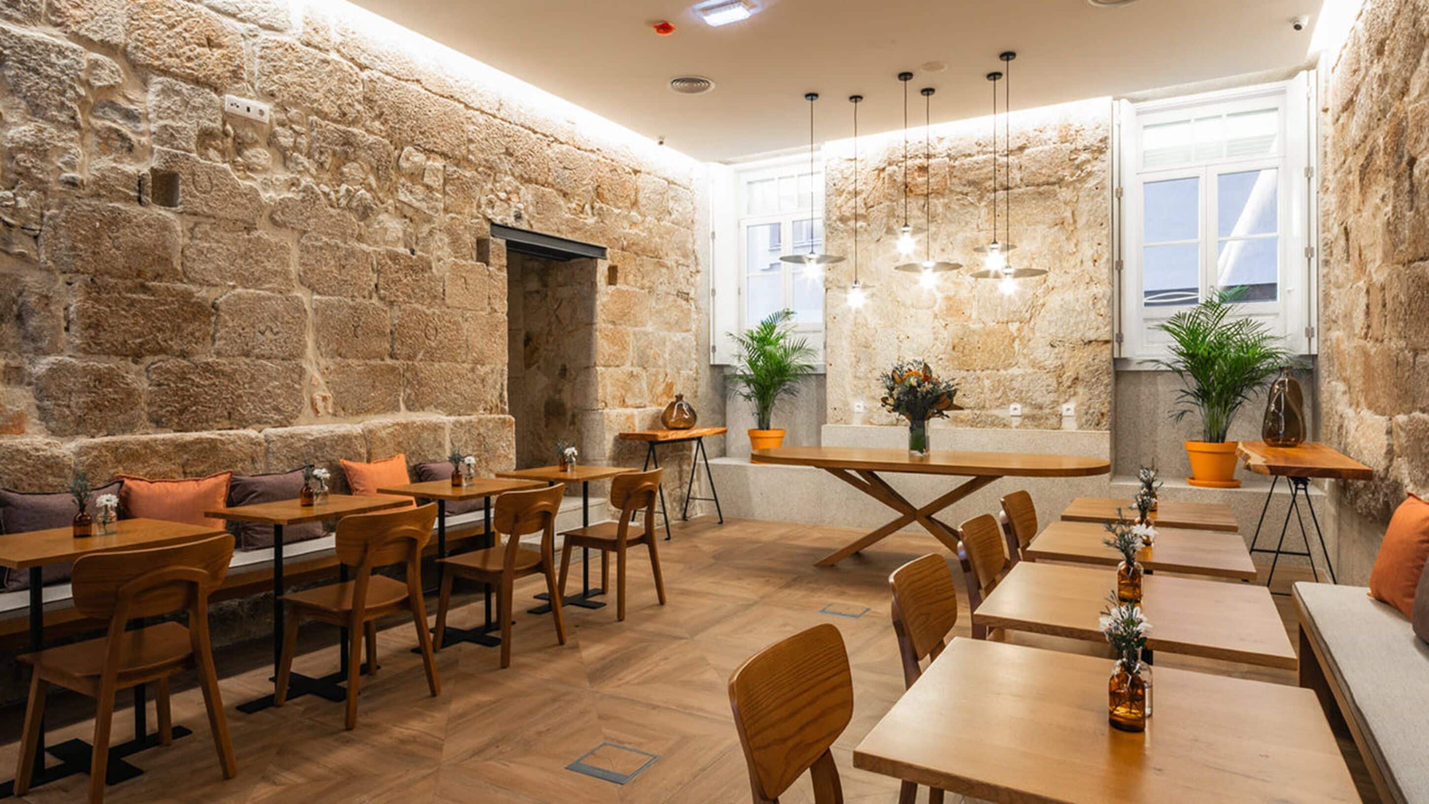 A cozy restaurant with stone walls, wooden tables, and chairs. There are potted plants and a large window.