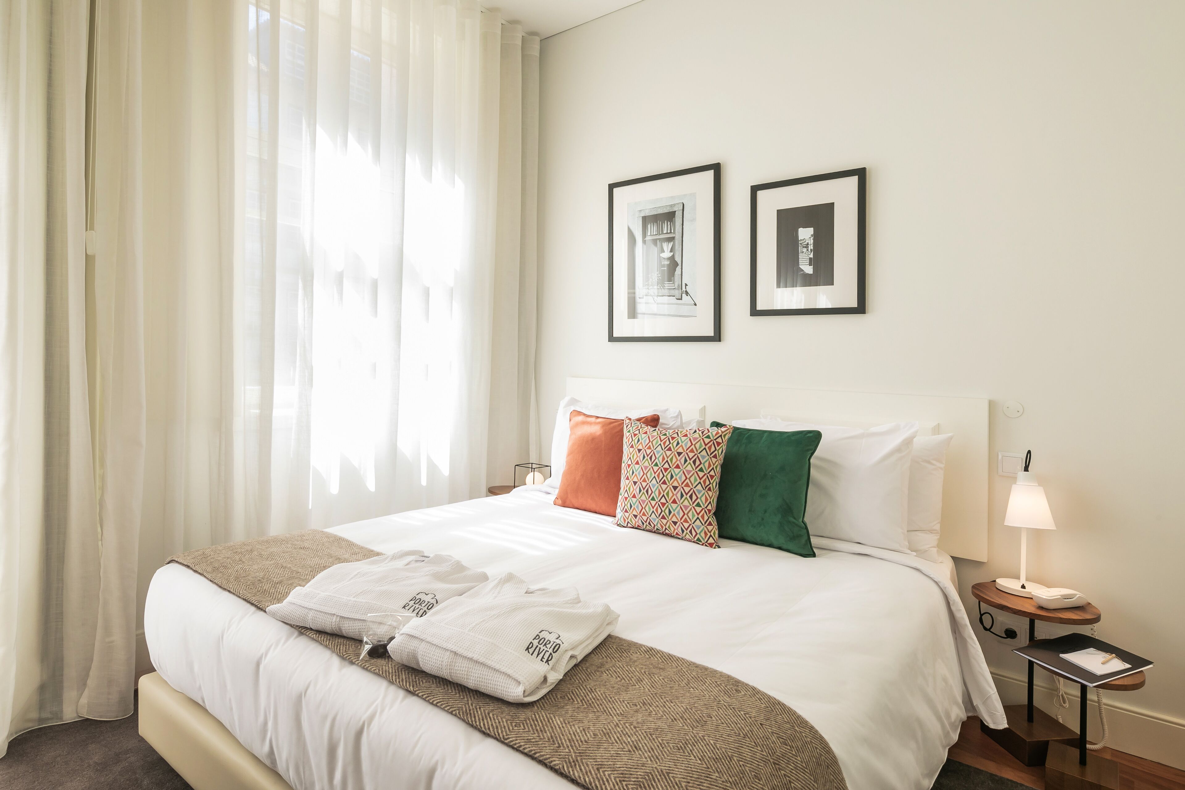 A neatly made bed with white linens, a brown throw blanket, and a set of towels with text. There are two pillows in different colors and two framed pictures on the wall. A bedside table with a lamp and a phone is visible.