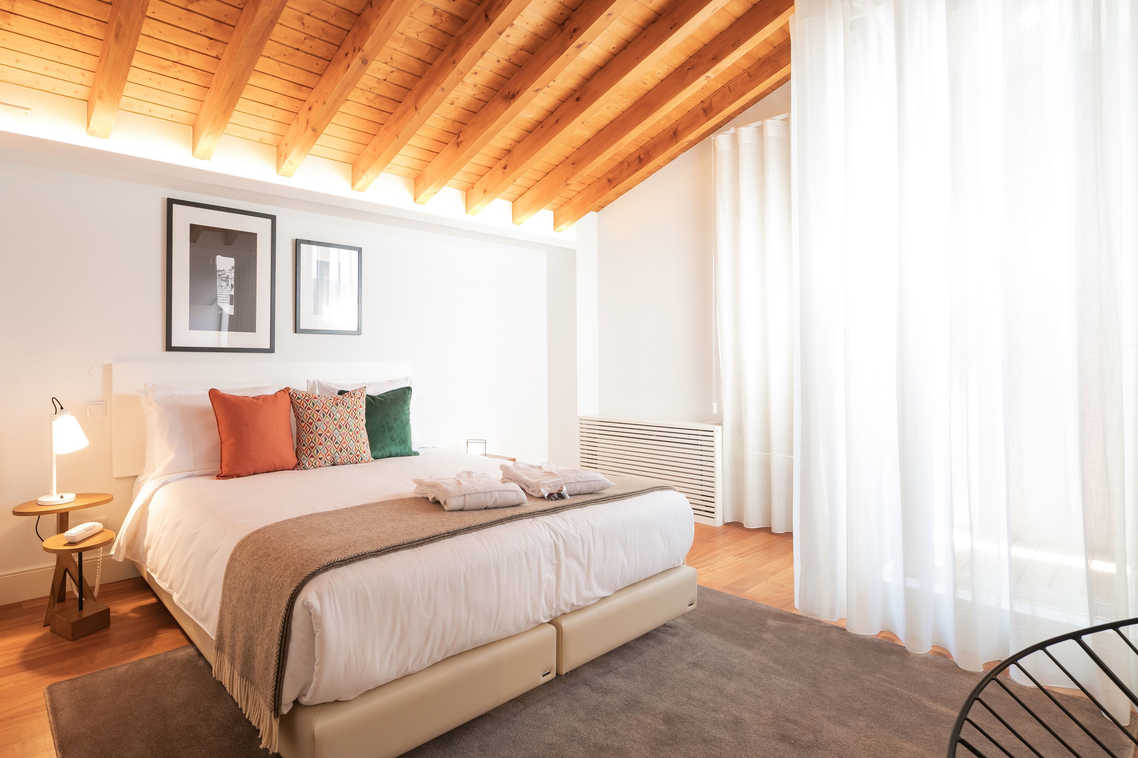 A bedroom with a wooden ceiling, a bed with white bedding, a nightstand with a lamp, and a window with sheer curtains.