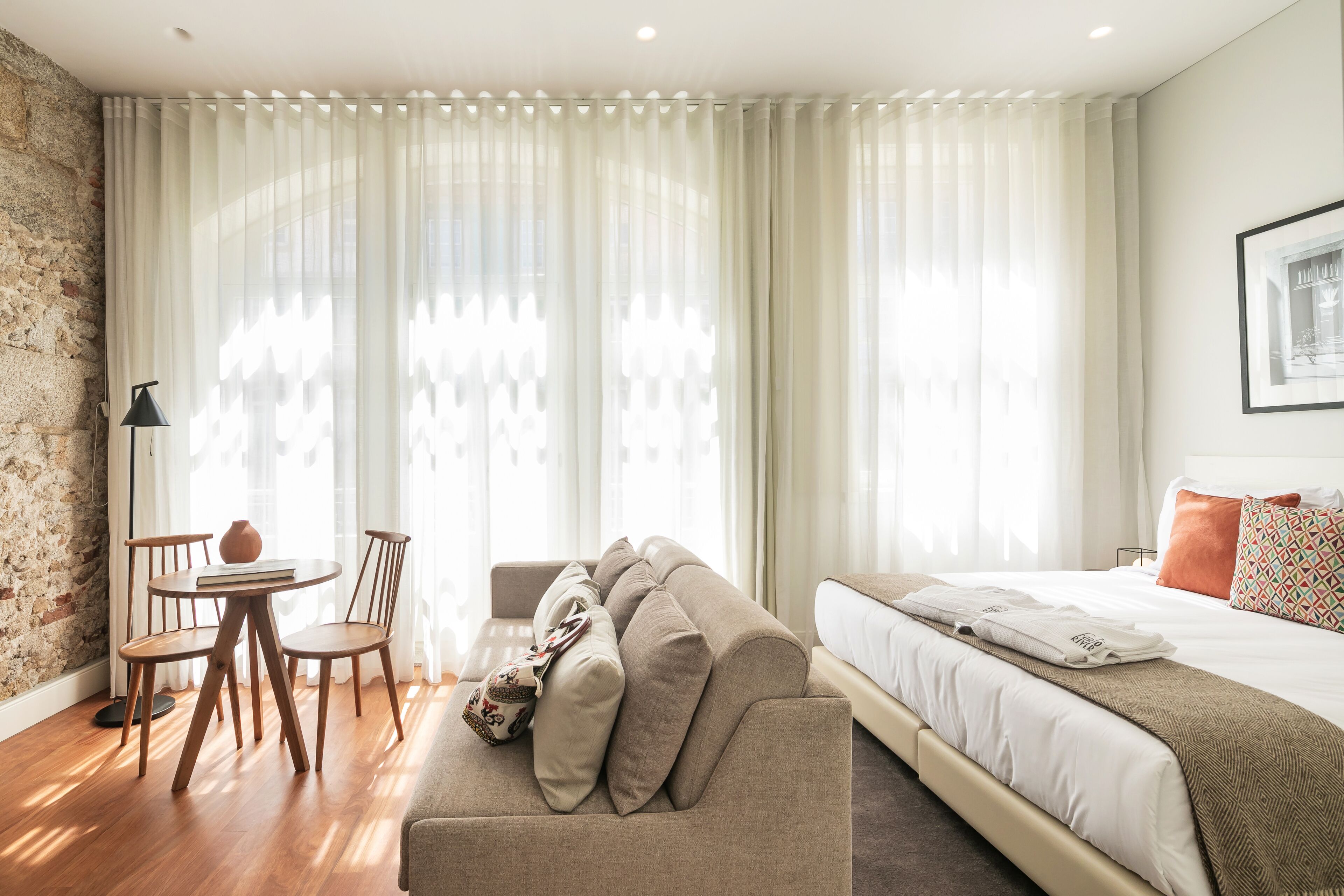 A modern hotel room with a large bed, a sofa, a dining table with chairs, and a stone wall.