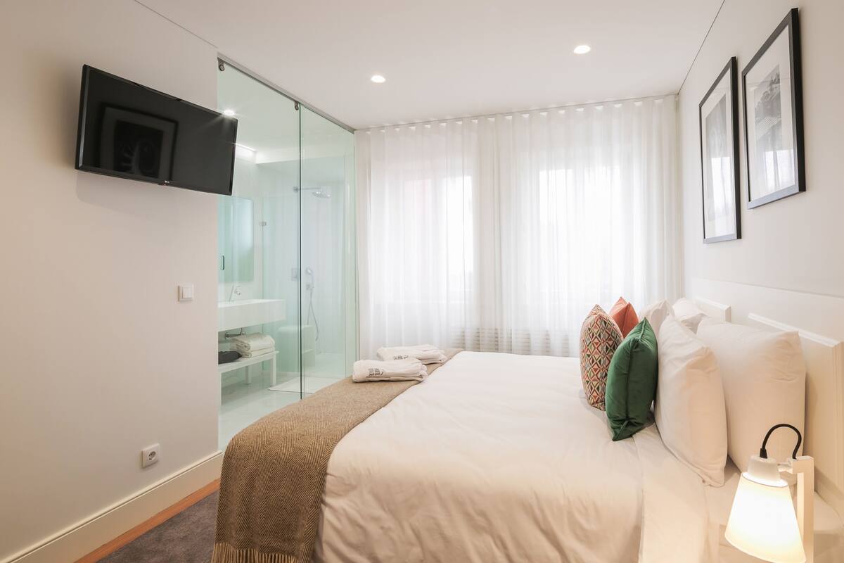Una habitación de hotel moderna con una cama grande, un televisor de pantalla plana montado en la pared, una cabina de ducha de vidrio y una mesita de noche con lámpara.