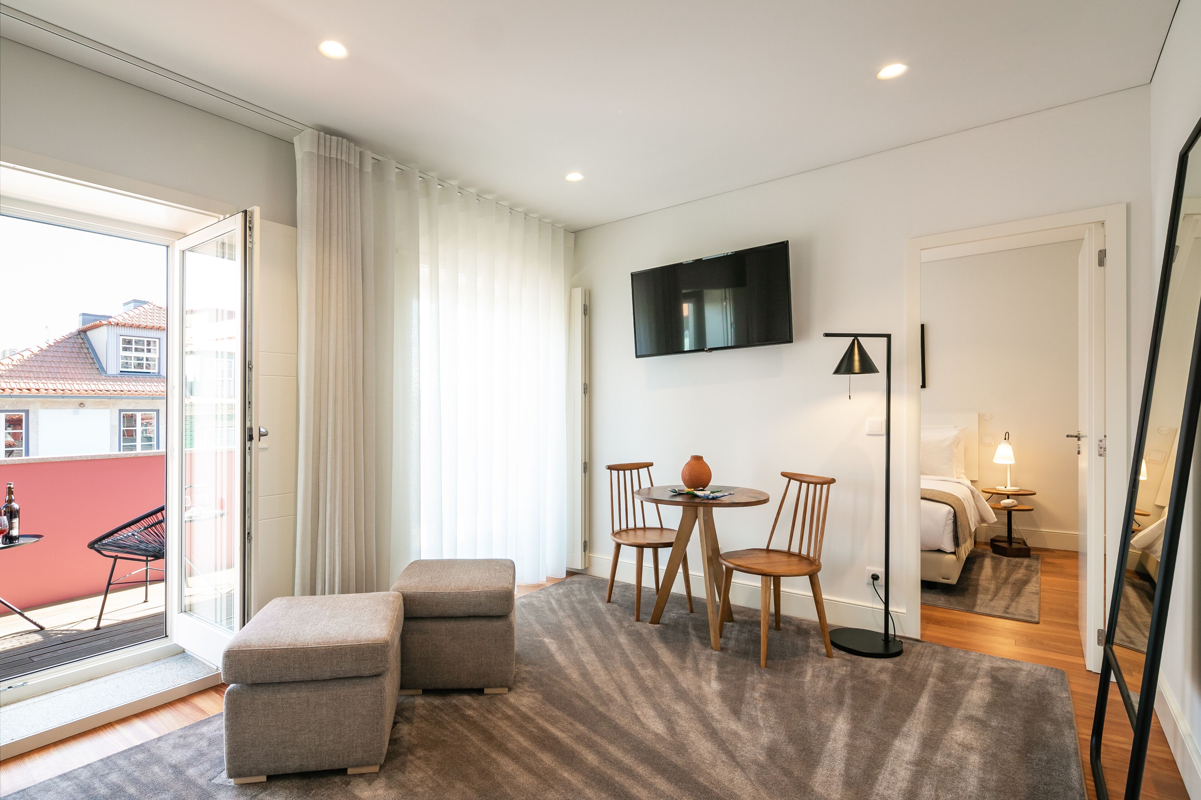 A modern hotel room with a sliding glass door to a balcony, a flat-screen TV mounted on the wall, a round dining table with chairs, a sofa, and a floor lamp.