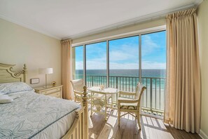 Habitación de hotel con cama, una pequeña mesa con sillas, una mesita de noche y vistas al océano a través de amplios ventanales.