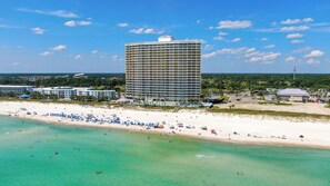 Un hotel frente a la playa con un edificio grande, una playa de arena y aguas turquesas transparentes.