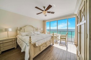 A bedroom with a large bed, a ceiling fan, a nightstand, a dresser, and a balcony with a view of the ocean.