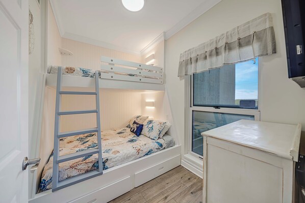 A bunk bed with a ladder, a window with curtains, a small cabinet, and a wall-mounted TV.