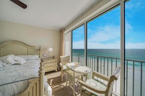 A bedroom with a bed, a nightstand, a chair, a table, and a large window with a view of the ocean.