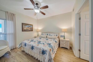 Un dormitorio con una cama, ventilador de techo, mesita de noche con lámpara, una silla de mimbre y una puerta.