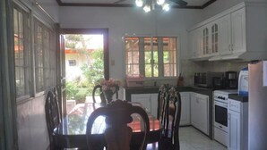 Una cucina con mobili bianchi, un tavolo da pranzo con sedie, una ventola a soffitto e una finestra vista sul verde.