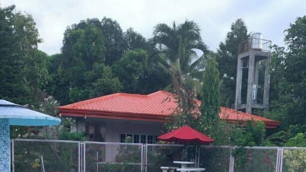 Una piscina con bordo piastrellato, area recintata e un edificio con tetto rosso sullo sfondo.