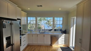 A modern kitchen with white cabinets, a stainless steel refrigerator, a dishwasher, and a large window providing a view of trees and a dock. - MOUNTAIN VIEWS fireplace SPA 5 bedrooms, air-cond, early checkin & late checkout (Wentworth Falls)
