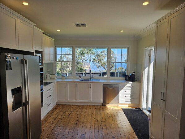 A modern kitchen with white cabinets, a stainless steel refrigerator, a dishwasher, and a large window providing a view of trees and a dock.