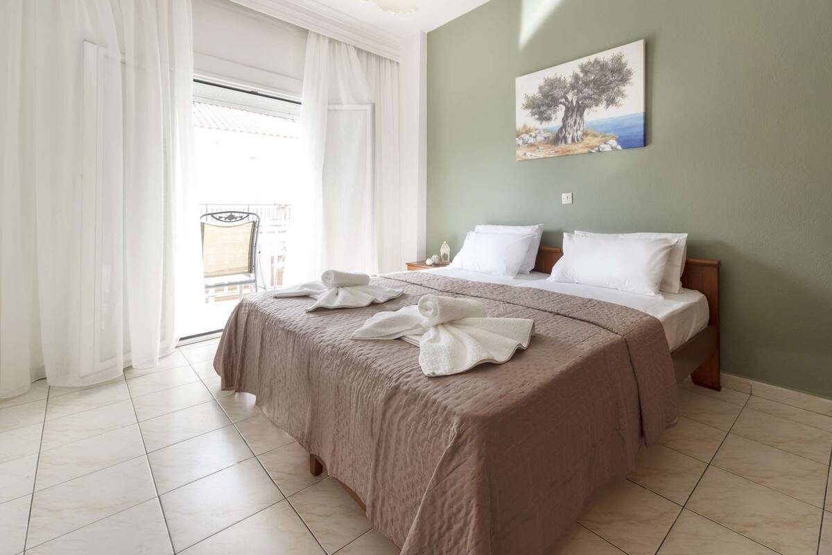 Ein Hotelzimmer mit einem großen Bett, weißer Bettwäsche, einem Gemälde an der Wand und Blick durch das Fenster.