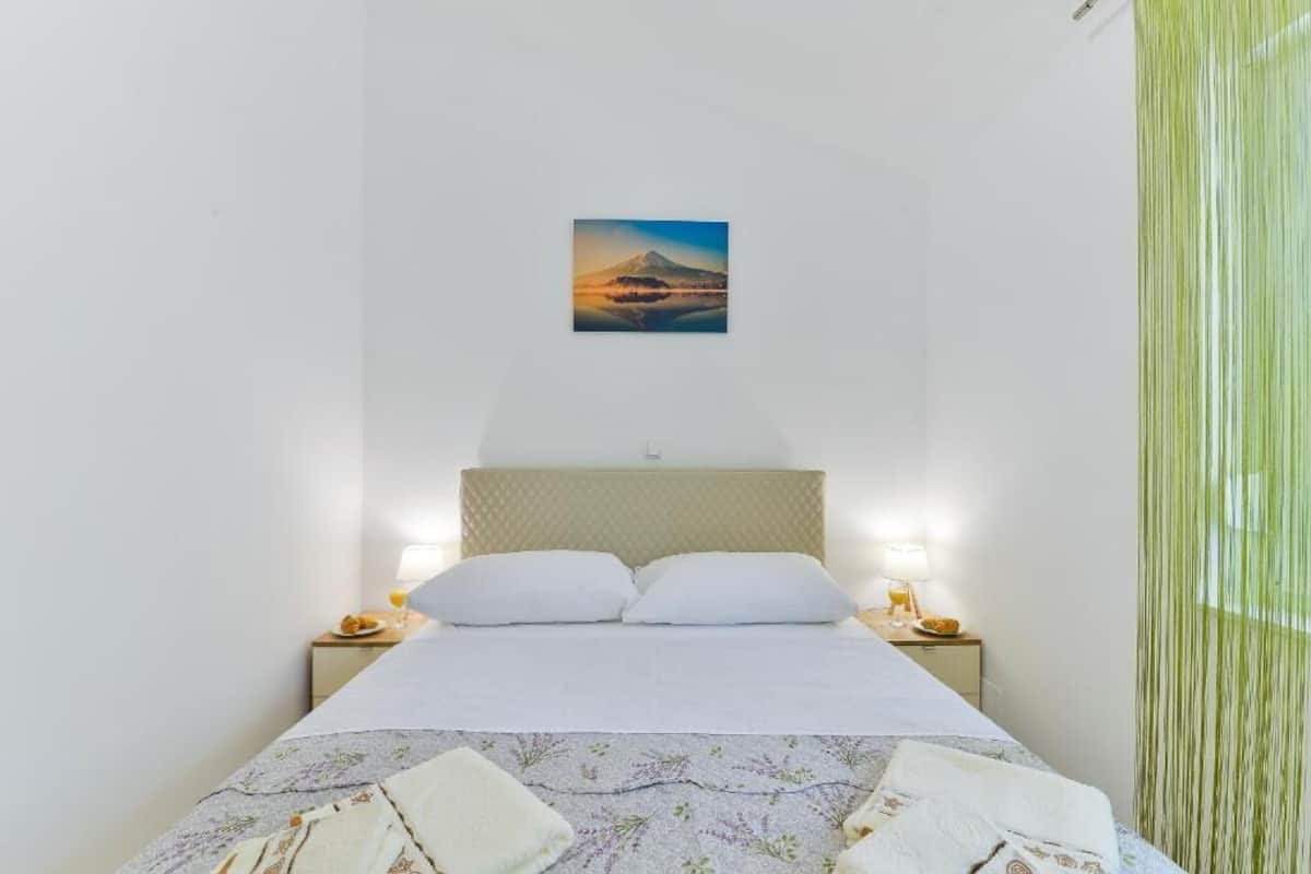 A hotel room with a bed, two bedside tables, a nightstand, a lamp, and a picture of a mountain on the wall.