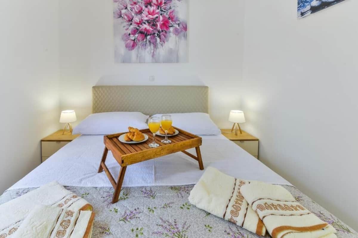 A neatly made bed with a wooden tray holding breakfast items, two bedside lamps, and a floral painting on the wall.