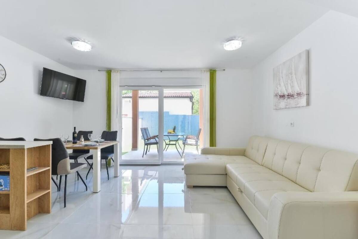 A modern living room with a sofa, a dining table with chairs, a TV mounted on the wall, and a sliding glass door leading to an outdoor area.