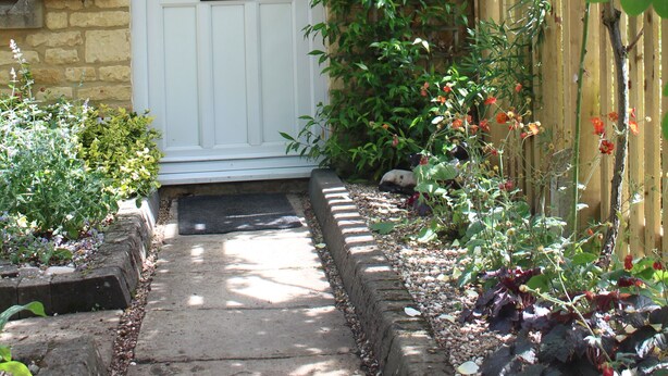 Een huisje met een witte deur, een bakstenen muur en een tuin met gele rozen en andere planten.