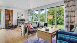 Una sala de estar acogedora con chimenea, un sofá, una mesa de comedor y ventanas amplias con vistas al jardín.