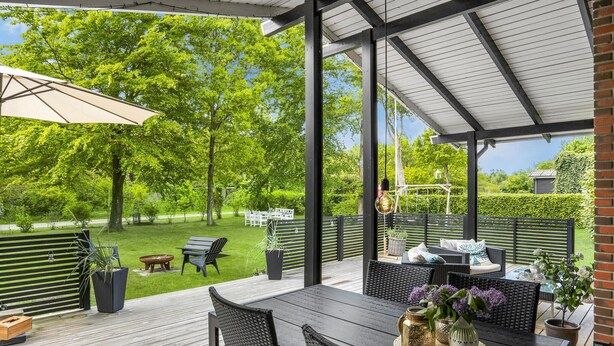 Un patio al aire libre con una mesa y sillas, rodeado por una terraza de madera y una zona cubierta con una pérgola.