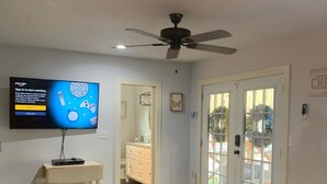 Una sala de estar con un sofá de cuero negro, un televisor de pantalla plana montado en la pared, un ventilador de techo y una puerta que da a un balcón.