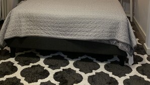 Un dormitorio ordenado con una cama, dos mesitas de noche con lámparas, un espejo redondo y un tapiz con textura en la pared.