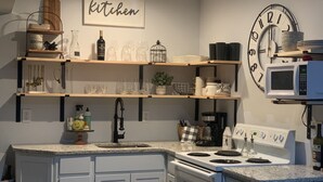 Una cocina moderna con armarios blancos, una estufa empotrada y un microondas. Hay estantes sobre la encimera con varios utensilios de cocina y un reloj en la pared.